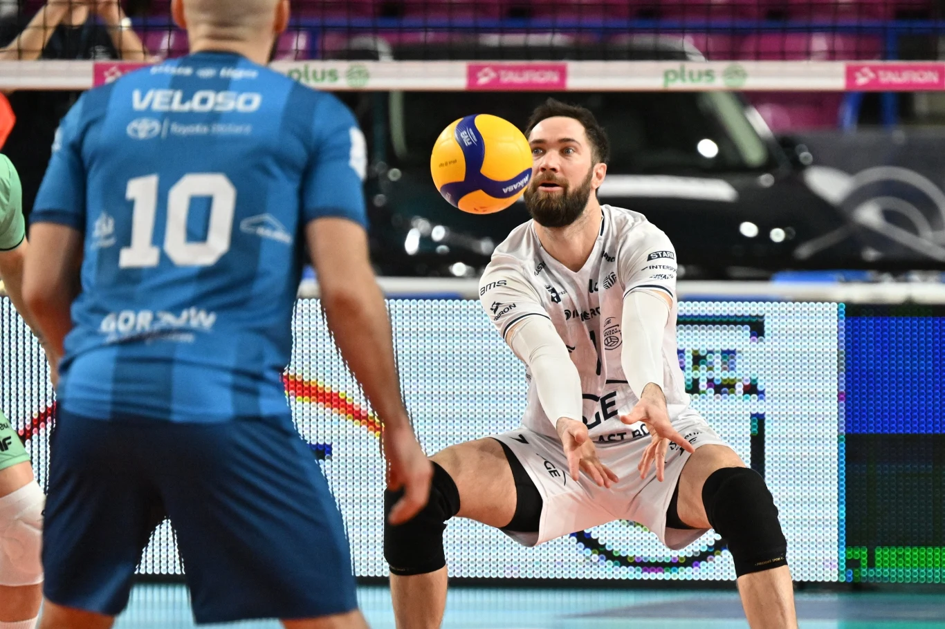 Zawodnik drużyny siatkarskiej w białym stroju odbija piłkę sposobem dolnym podczas meczu, w tle widoczny zawodnik drużyny przeciwnej w niebieskim stroju z numerem 10. Zawodnik drużyny siatkarskiej w białym stroju odbija piłkę sposobem dolnym podczas meczu, w tle widoczny zawodnik drużyny przeciwnej w niebieskim stroju z numerem 10.