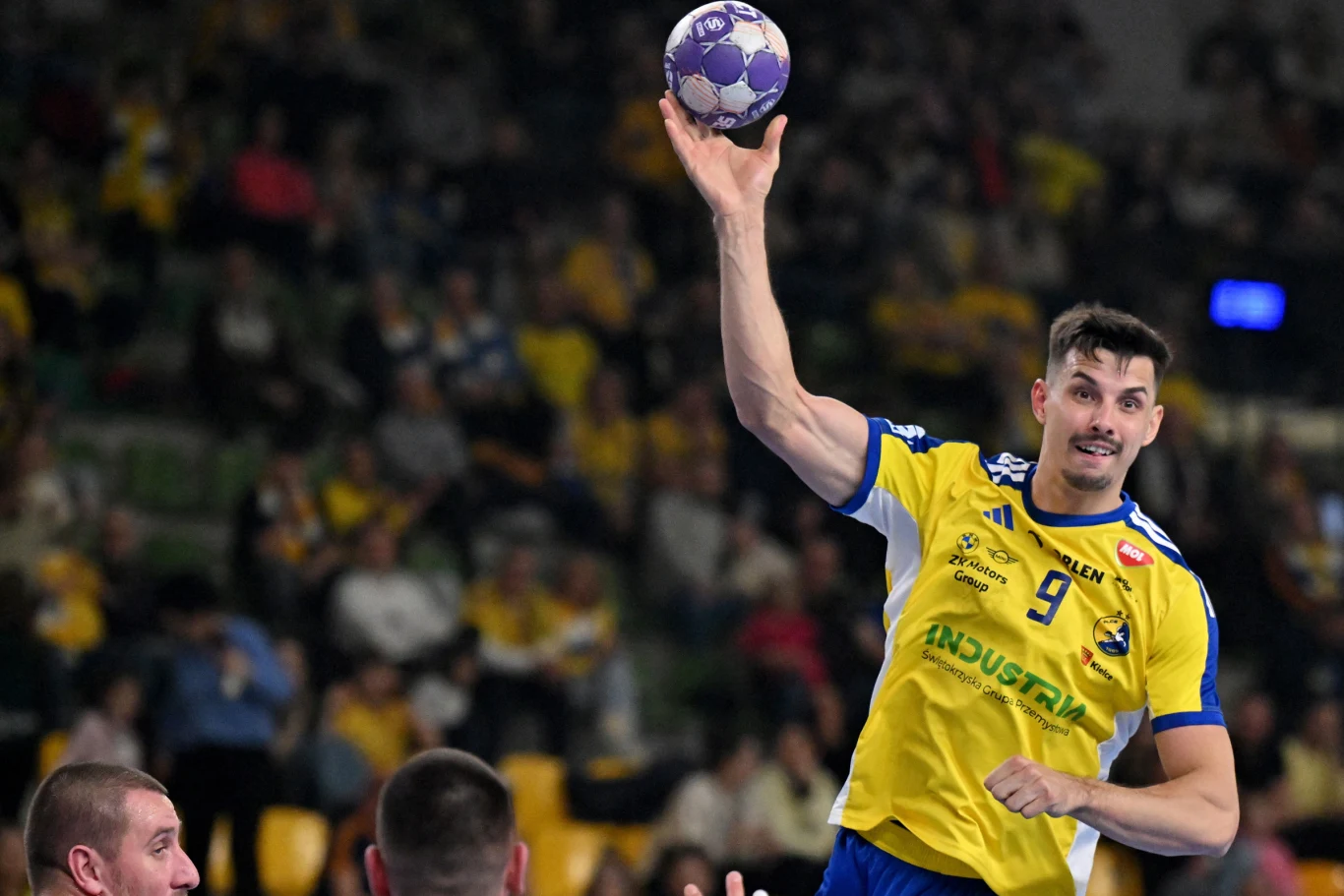 Zawodnik ubrany w żółto-niebieski strój sportowy unosi rękę z piłką do rzutu podczas meczu piłki ręcznej, na drugim planie widoczna jest publiczność na trybunach. Zawodnik ubrany w żółto-niebieski strój sportowy unosi rękę z piłką do rzutu podczas meczu piłki ręcznej, na drugim planie widoczna jest publiczność na trybunach.