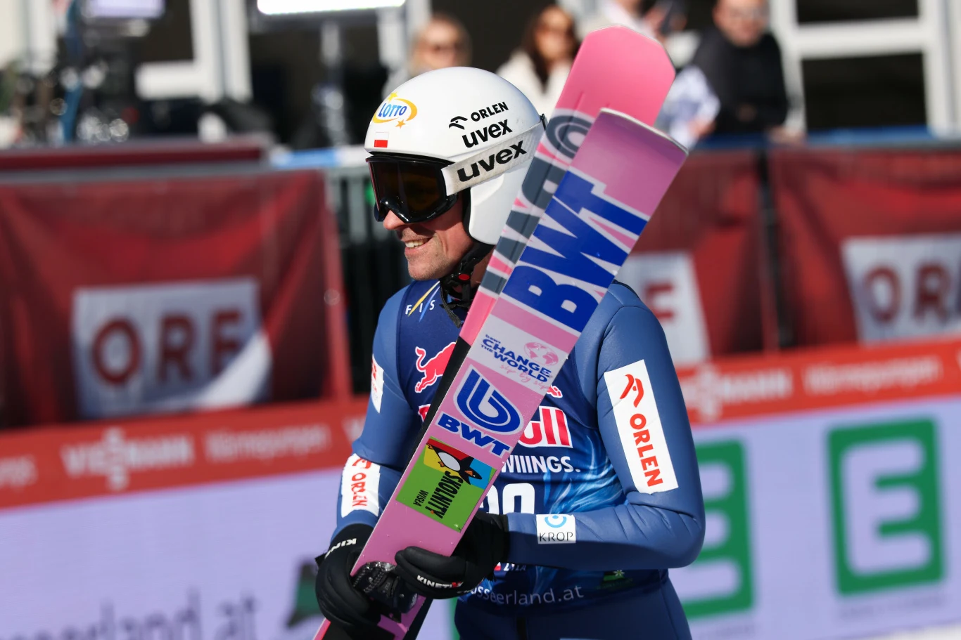 Skoczek narciarski w niebieskim kombinezonie oraz kasku ochronnym trzyma w rękach charakterystyczne narty z widocznymi sponsorami, stojąc na tle band z reklamami przy skoczni.