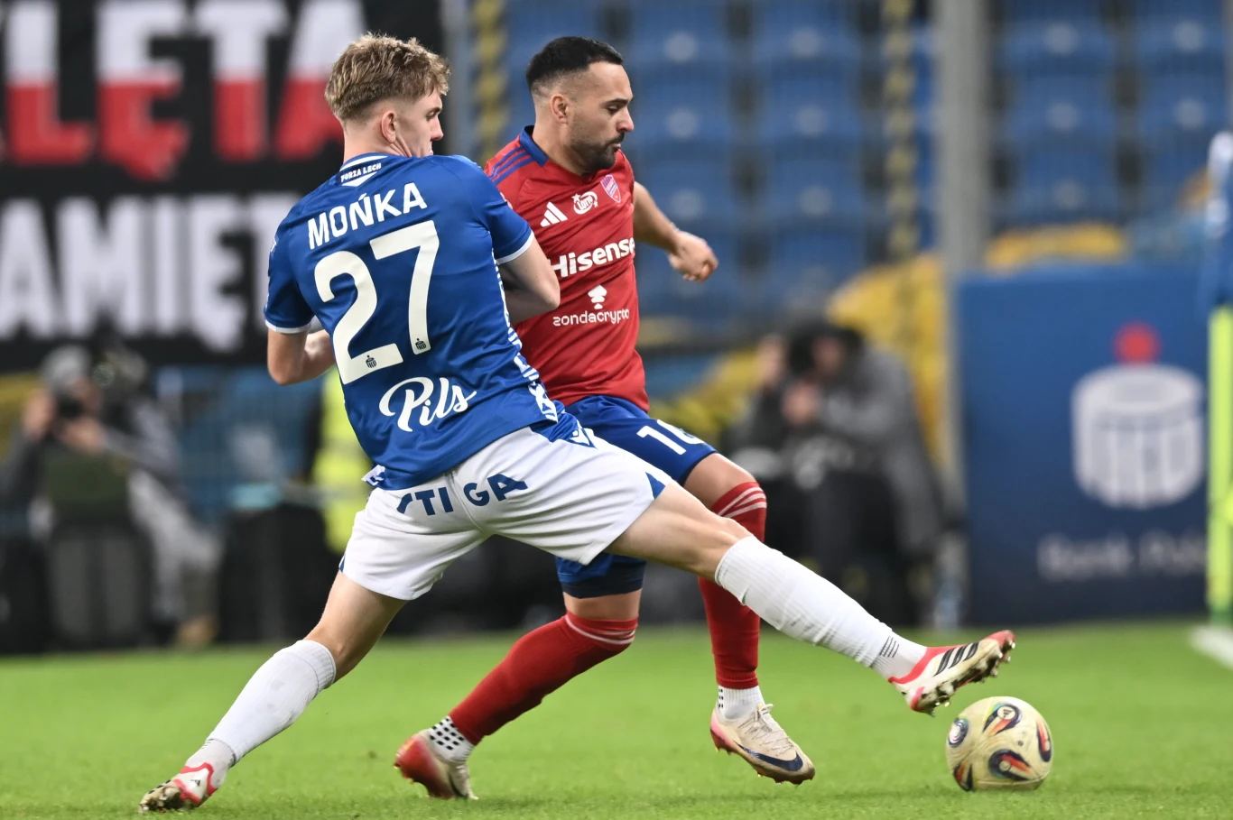 Dwóch piłkarzy walczy o piłkę podczas meczu piłkarskiego na stadionie, jeden ubrany w niebieską koszulkę z numerem 27, drugi w czerwoną koszulkę, intensywna rywalizacja na tle trybun. Dwóch piłkarzy walczy o piłkę podczas meczu piłkarskiego na stadionie, jeden ubrany w niebieską koszulkę z numerem 27, drugi w czerwoną koszulkę, intensywna rywalizacja na tle trybun.