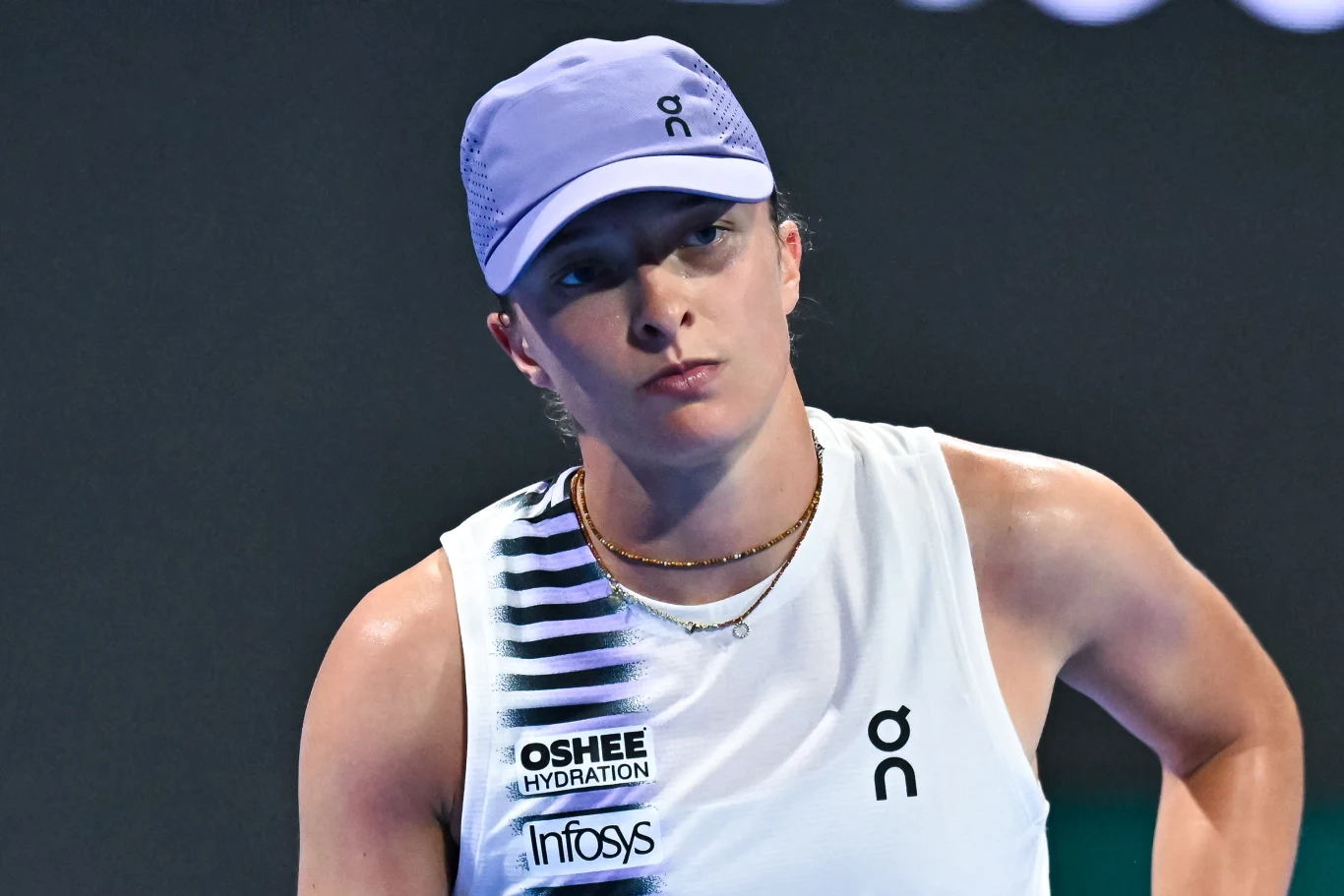 Tenisistka w białym stroju sportowym i fioletowej czapce z daszkiem, z wyraźnym skupieniem na twarzy, podczas meczu na oświetlonym korcie.