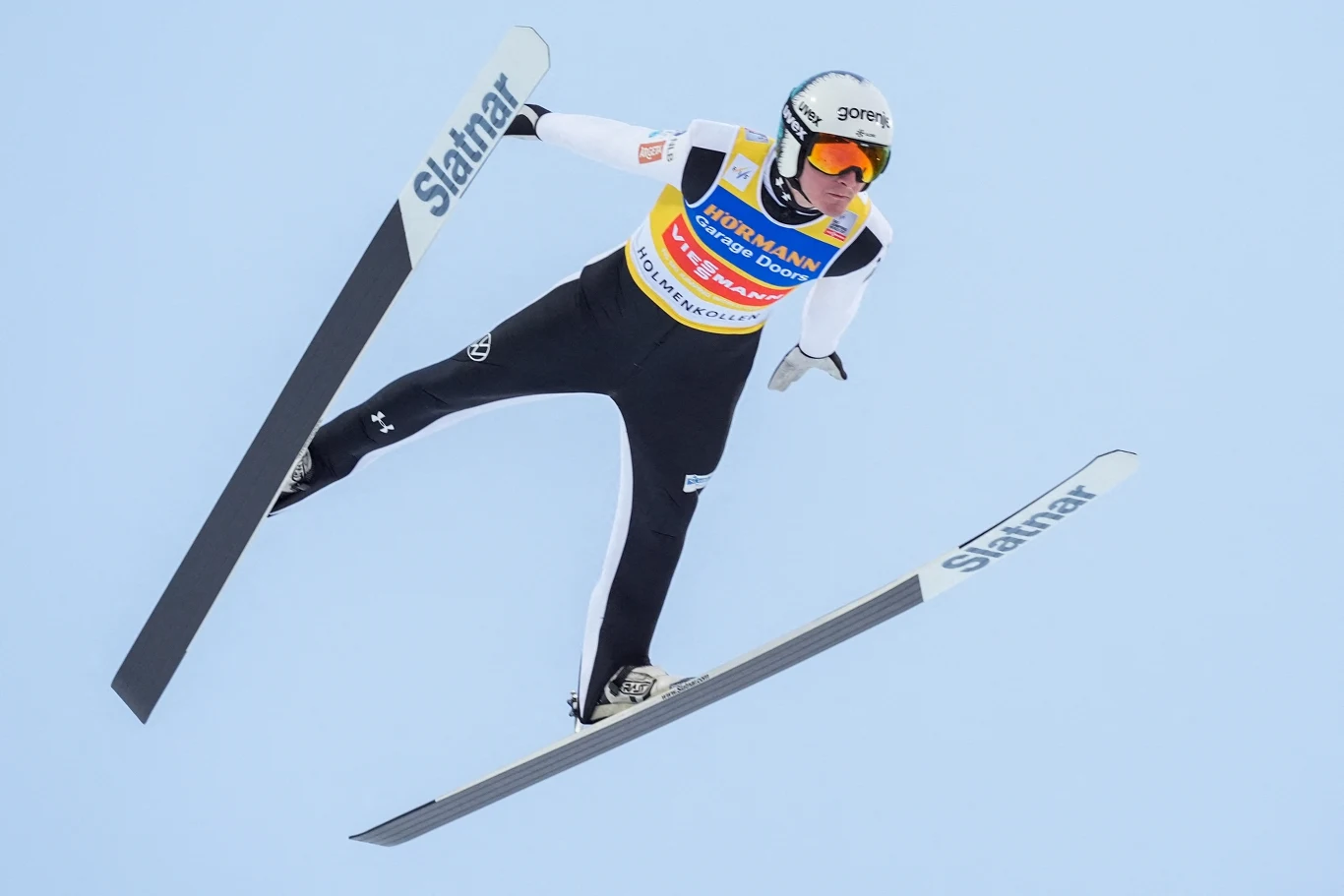 skoczek narciarski w locie na tle jasnego nieba, wyprostowane narty ułożone w kształcie litery V, sportowy kombinezon i kask z okularami ochronnymi, widoczne logo sponsora na piersi zawodnika