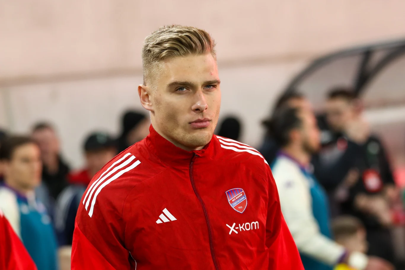 młody mężczyzna w czerwonej sportowej kurtce z herbem klubu i logo sponsora, z poważnym wyrazem twarzy, na tle innych osób w strojach sportowych na ławce rezerwowych