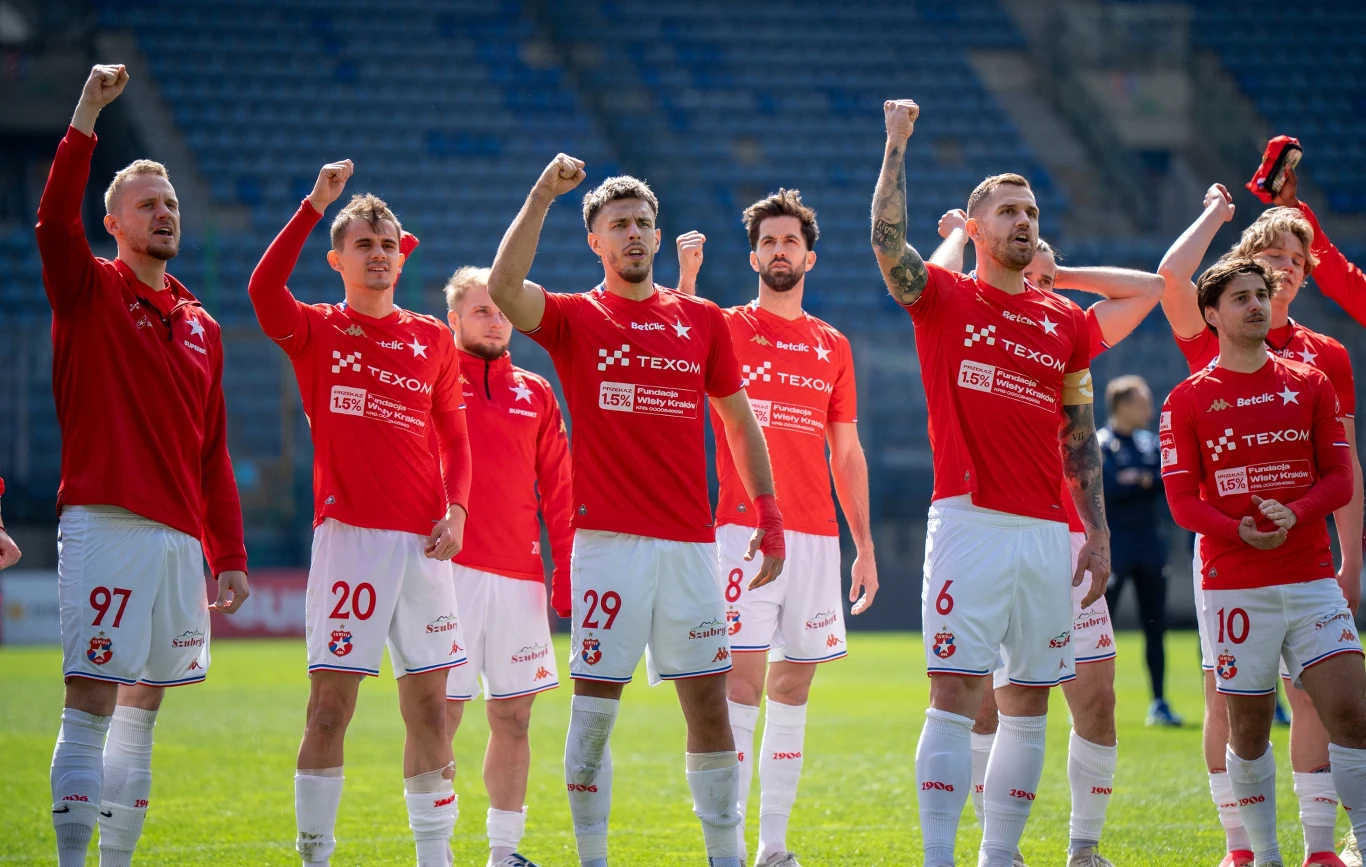 grupa piłkarzy w czerwonych koszulkach i białych spodenkach stoi na murawie stadionu z uniesionymi w geście triumfu pięściami, tworząc zgrany zespół, który celebruje wspólny sukces