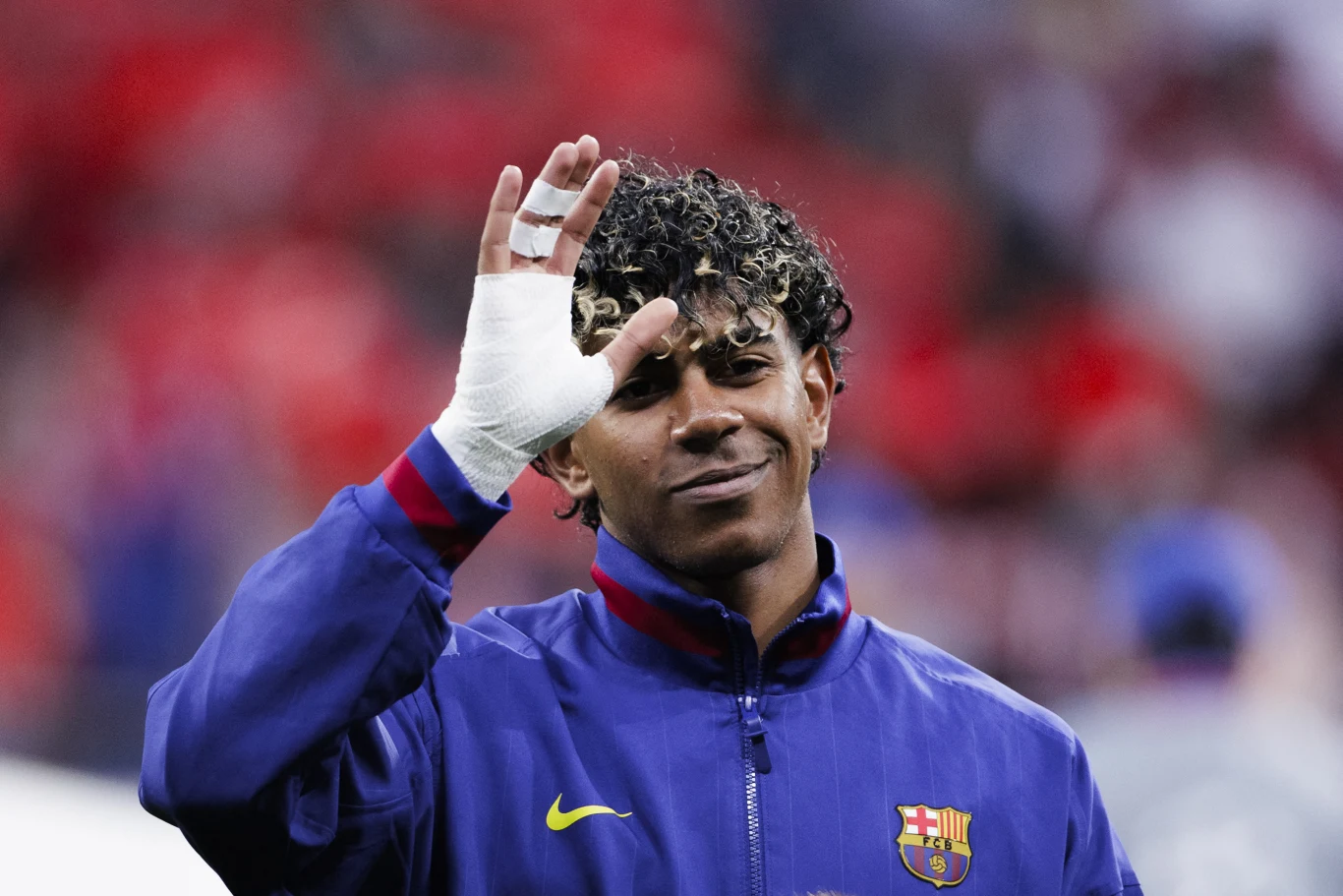 piłkarz w niebieskiej kurtce FC Barcelona wykonuje gest dłonią, na której ma opatrunek bandażowy, na tle rozmytych, czerwonych barw trybun stadionu