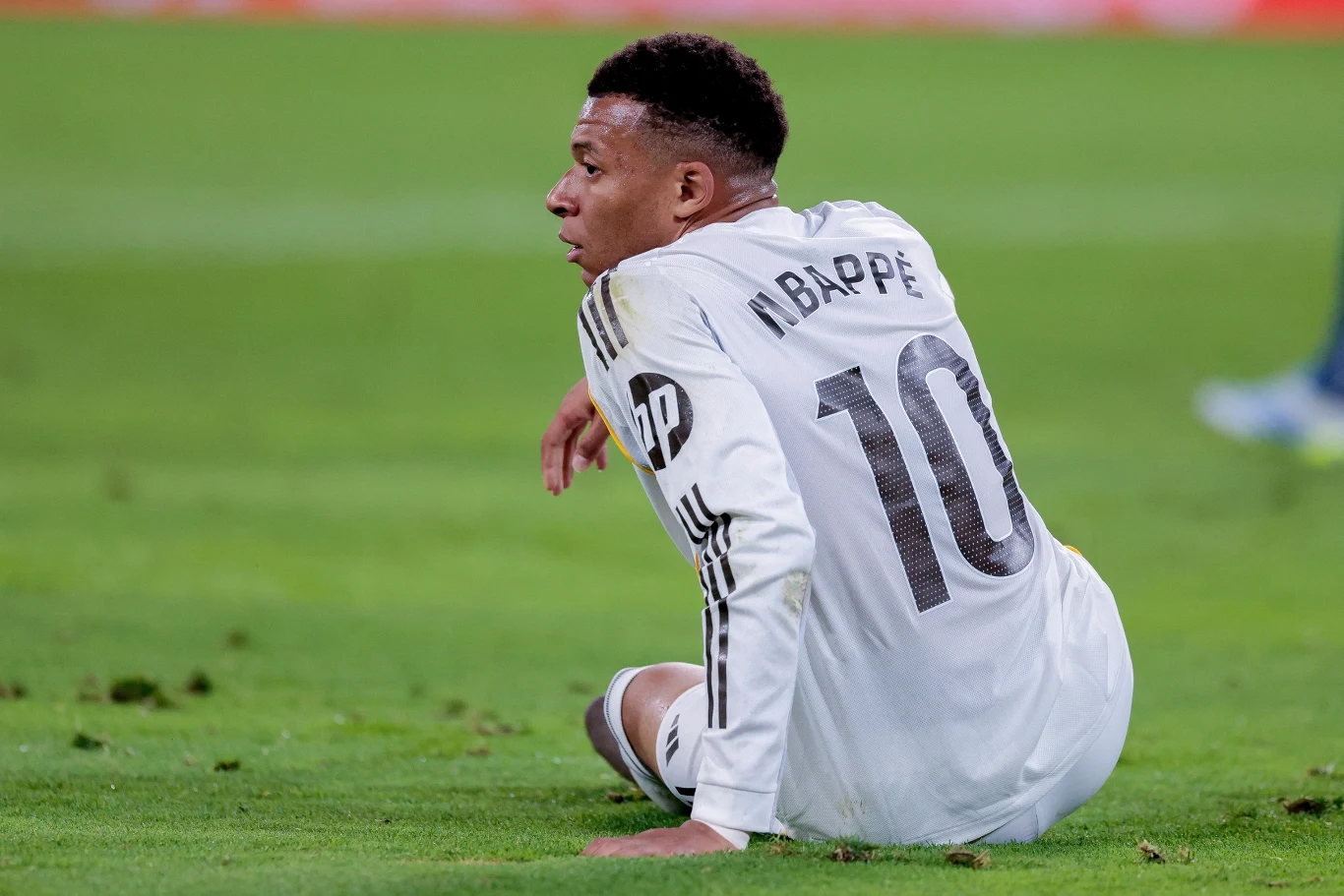 Zawodnik w białym stroju piłkarskim z numerem 10 i nazwiskiem „Mbappé” siedzi na murawie stadionu, spoglądając w bok.