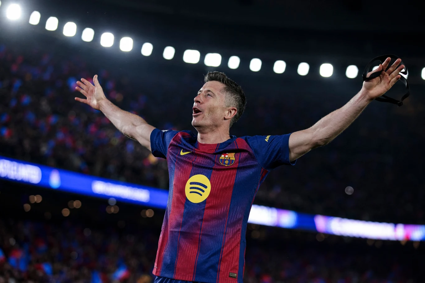 Piłkarz w koszulce FC Barcelony z szeroko rozłożonymi ramionami świętuje na stadionie oświetlonym jupiterami.