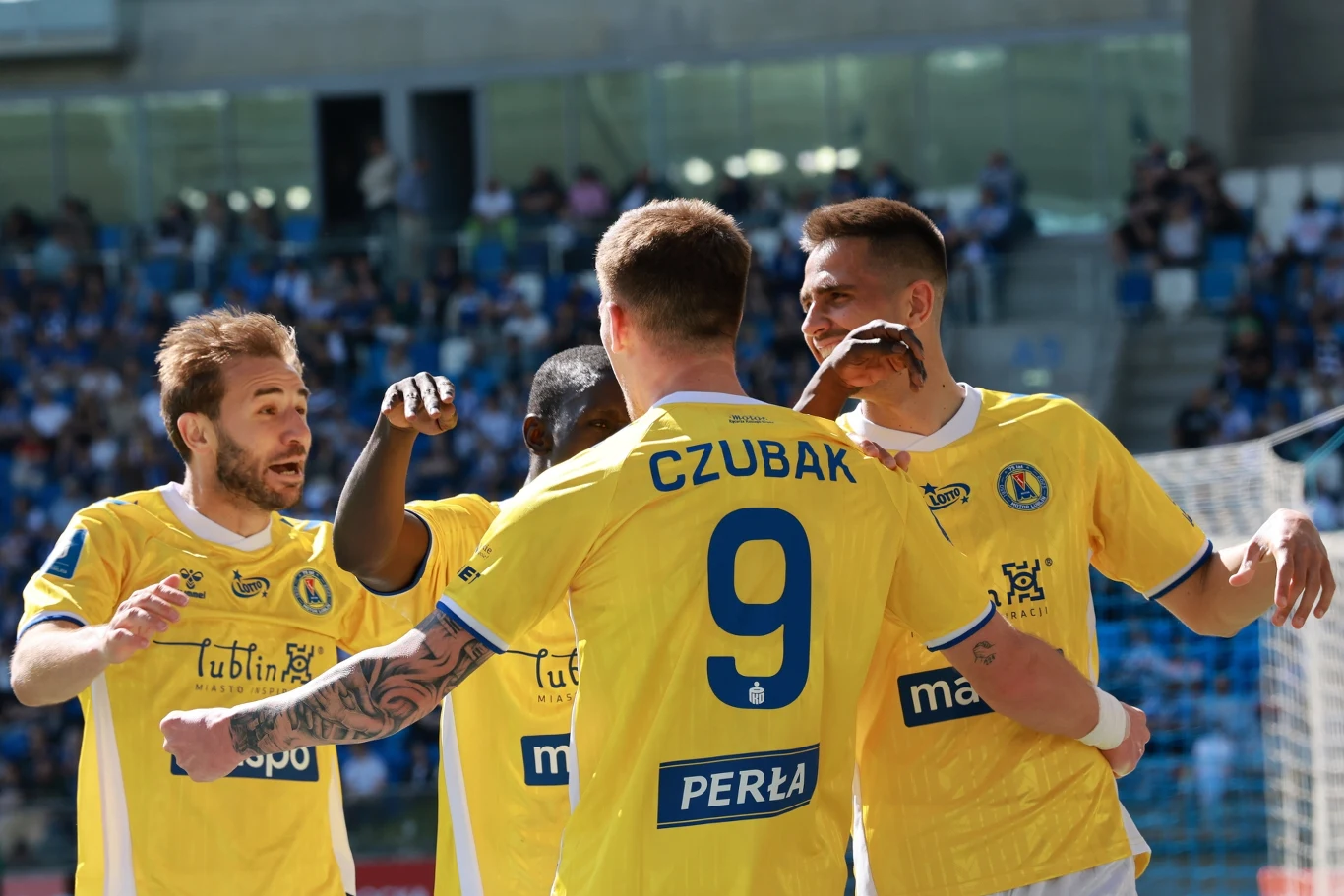 Piłkarze w żółtych koszulkach świętują zdobycie gola podczas meczu na stadionie, skupieni wokół zawodnika z numerem 9.