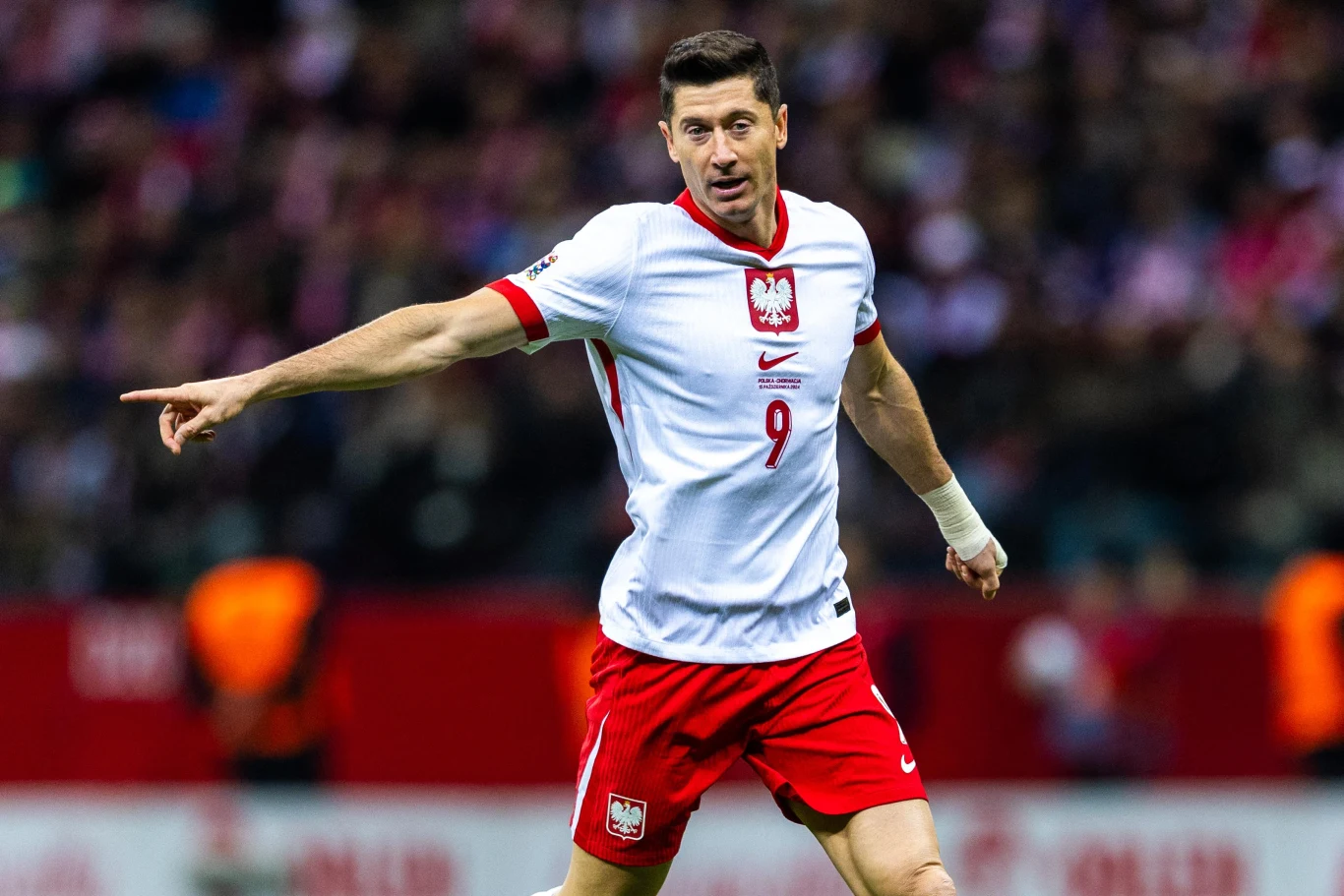 Robert Lewandowski Piłkarz w biało-czerwonym stroju z numerem 9 wskazuje ręką kierunek podczas meczu na stadionie.