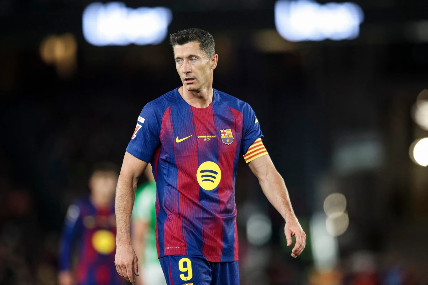 Piłkarz w koszulce FC Barcelony z opaską kapitańską na ramieniu, stojący na murawie stadionu podczas meczu.