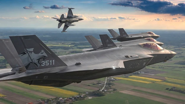 Dwa nowoczesne myśliwce F-35 lecące w formacji na niewielkiej wysokości nad polami, w tle wyżej widoczny myśliwiec F-16.