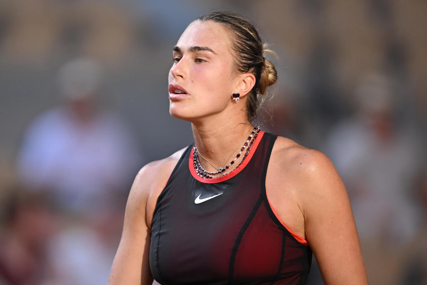 Kobieta w sportowym stroju z ciemnym topem i czerwonymi akcentami, zapiętymi włosami oraz naszyjnikami, w lekkim makijażu, wygląda na skoncentrowaną i zmęczoną po wysiłku fizycznym.