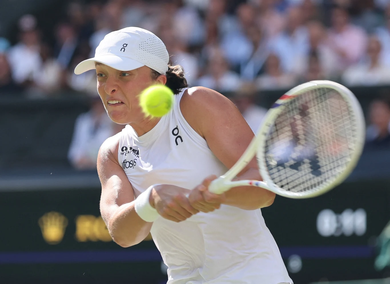 Tenisistka w białym stroju wykonuje dwuręczny backhand, skupiona na lecącej w jej kierunku piłce.