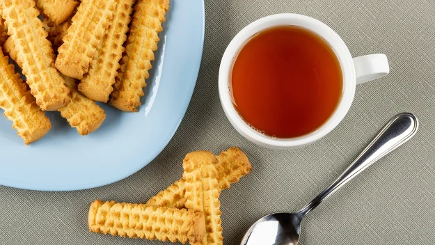 Herbata w białej filiżance, ciastka maślane na niebieskim talerzu oraz srebrna łyżeczka na szarej powierzchni. Herbata w białej filiżance, ciastka maślane na niebieskim talerzu oraz srebrna łyżeczka na szarej powierzchni.