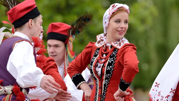 Grupa młodych ludzi w tradycyjnych polskich strojach ludowych wykonuje taniec ludowy, kobieta ubrana w haftowany czerwony strój i biały czepiec uśmiecha się, a mężczyźni mają charakterystyczne czerwone czapki z piórami.