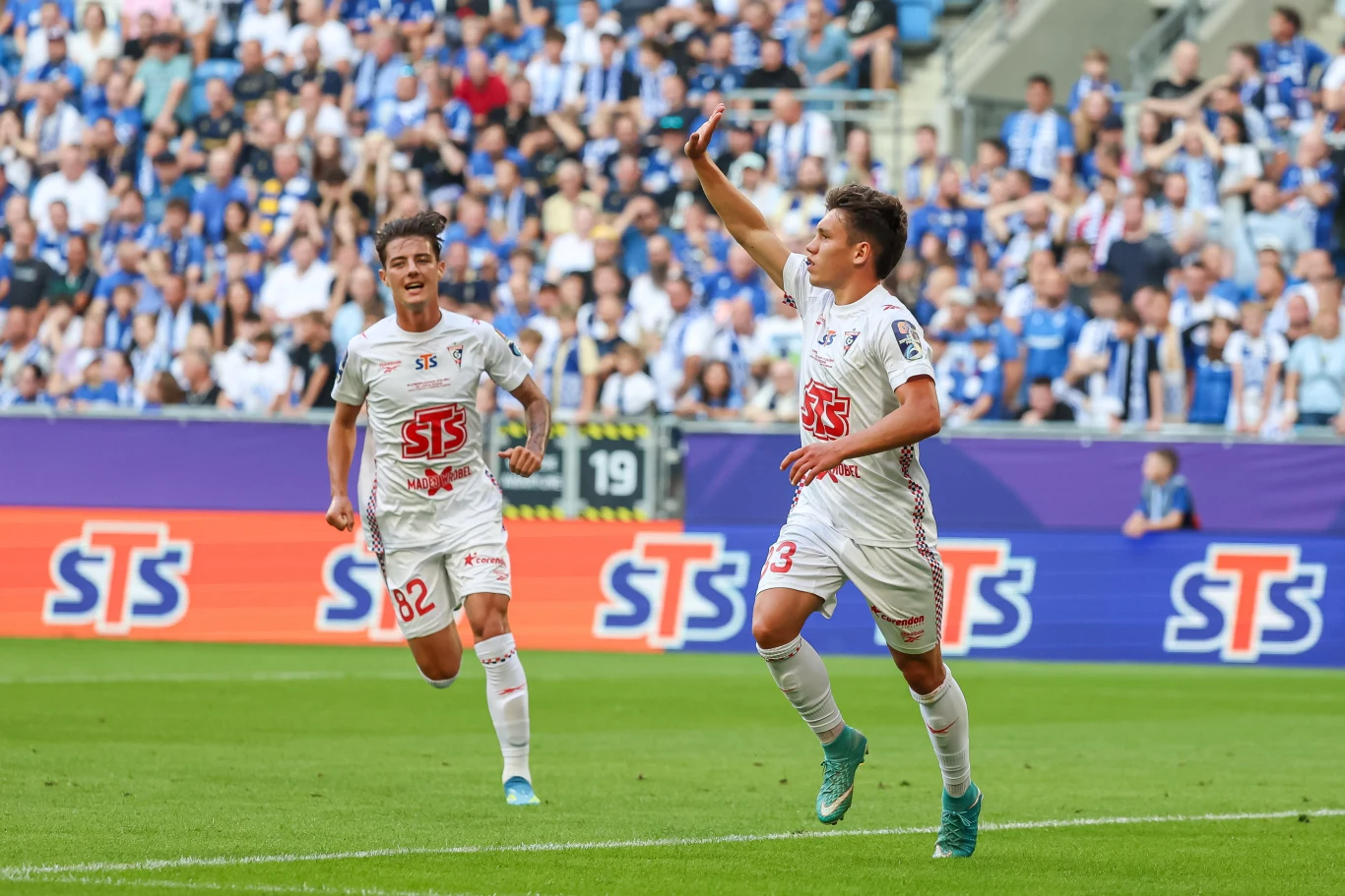 Dwóch piłkarzy w białych strojach świętuje zdobycie gola na stadionie pełnym kibiców, jeden z zawodników unosi rękę w geście triumfu.