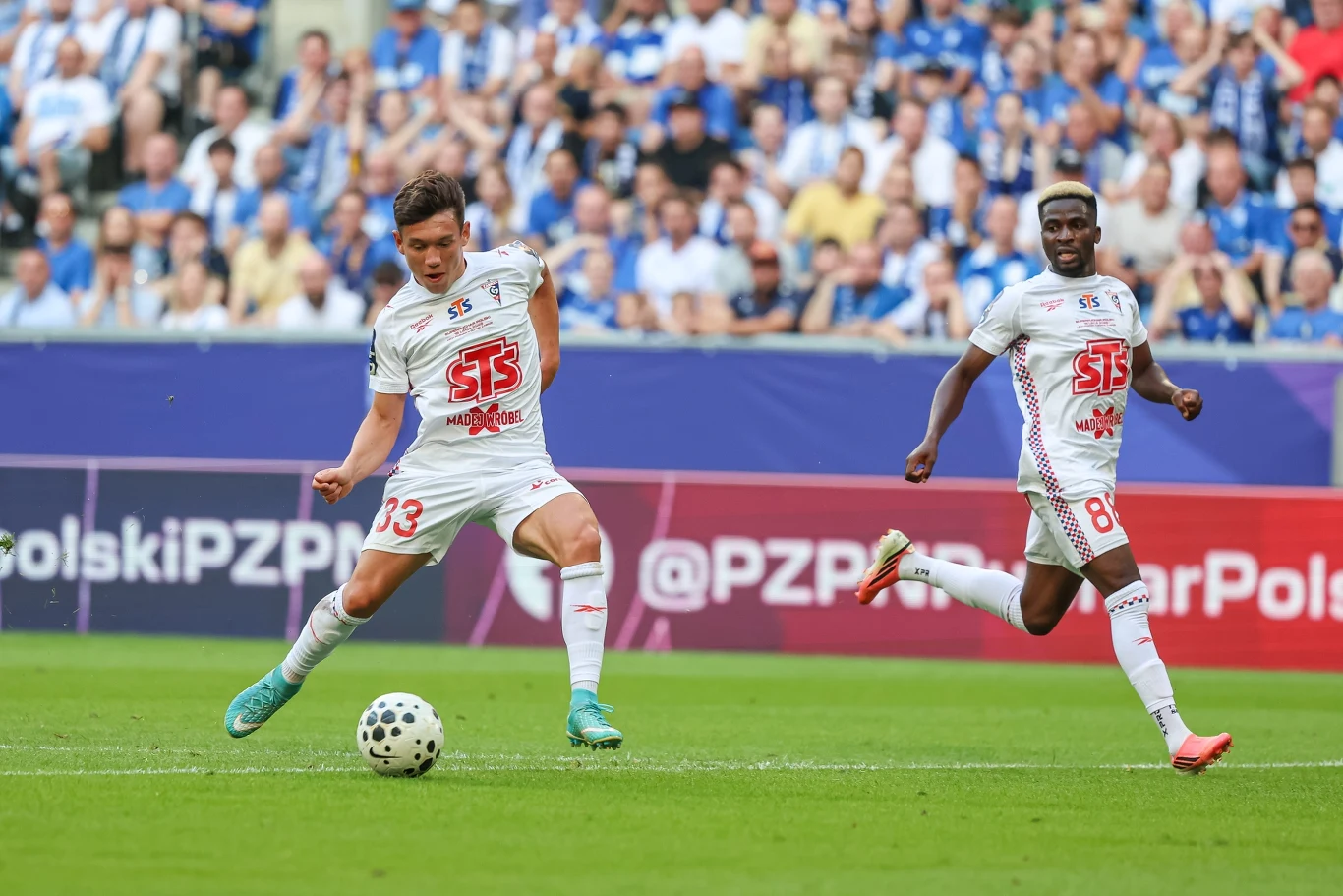 Dwóch piłkarzy w białych strojach z czerwonymi numerami i logo sponsora, jeden z nich kopie piłkę podczas meczu na zielonym boisku, w tle widoczni kibice na trybunach.