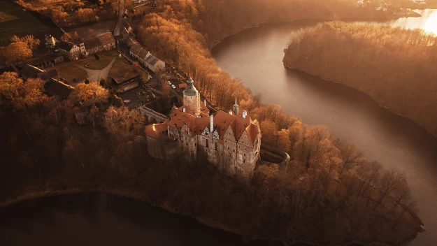 Zamek z czerwonym dachem otoczony lasem na cyplu, wokół spokojna woda rzeki tworząca naturalną granicę.