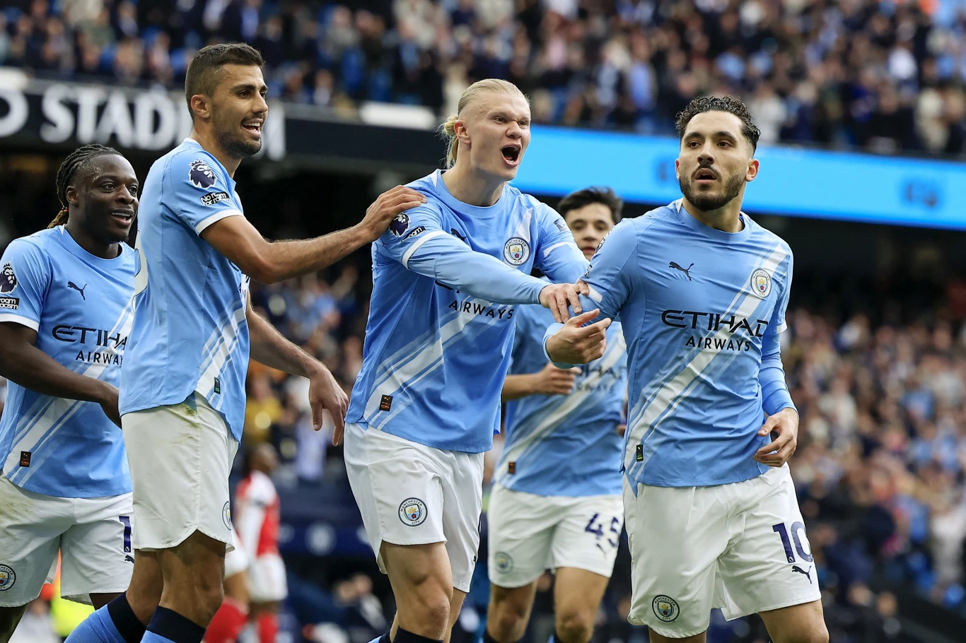 Piłkarze Manchesteru City w błękitnych koszulkach świętują gola na murawie, wyrażając emocje i radość w trakcie meczu przed trybunami pełnymi kibiców.
