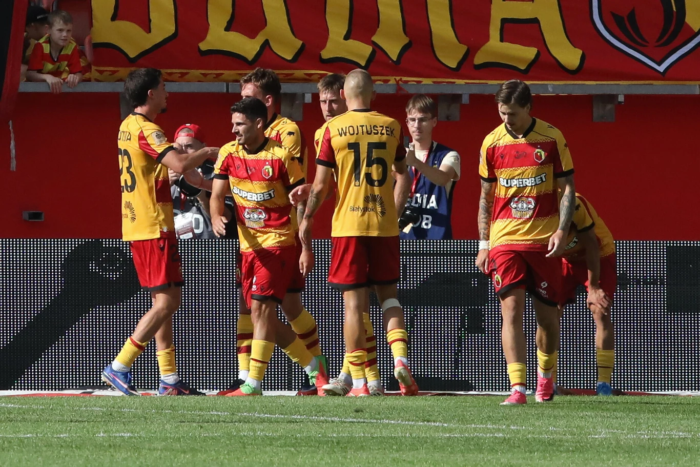 Grupa piłkarzy w żółto-czerwonych strojach świętuje razem na boisku, wokół nich widoczne są sportowe banery i fragmenty stadionu.