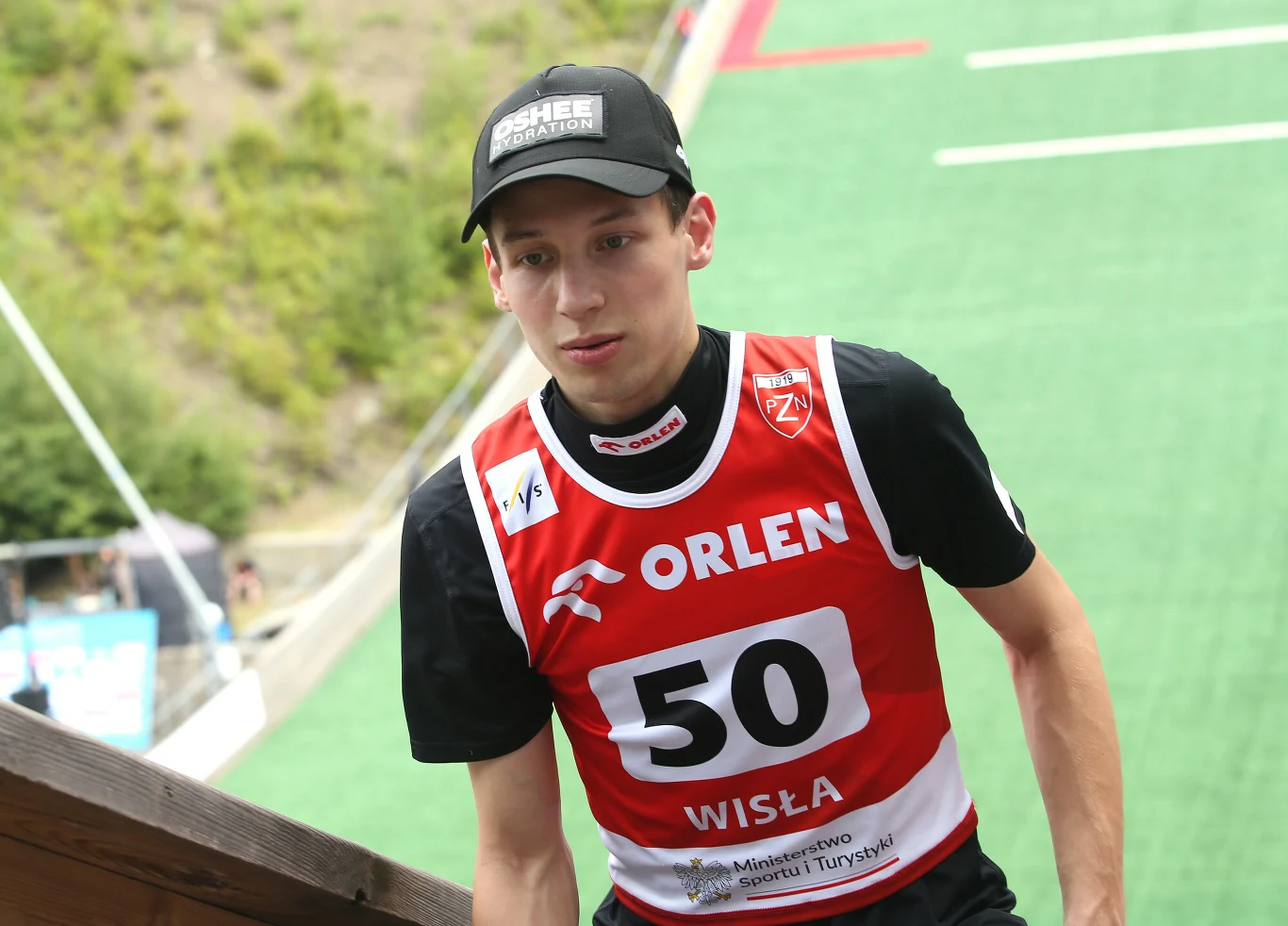 Zawodnik w sportowym stroju z numerem 50 oraz czarną czapką idzie po schodach podczas zawodów skoków narciarskich na tle zielonej skoczni w Wiśle.