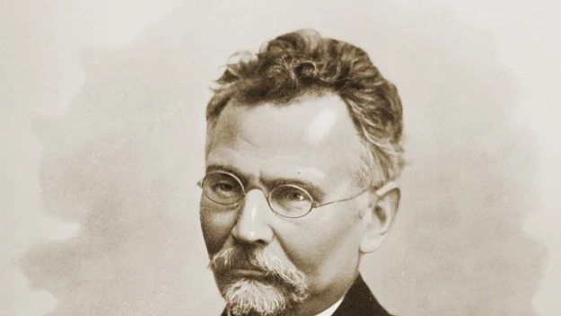 Portret mężczyzny z siwiejącymi włosami i brodą, ubrany w ciemny garnitur, białą koszulę oraz okulary, o poważnym wyrazie twarzy. Portret mężczyzny z siwiejącymi włosami i brodą, ubrany w ciemny garnitur, białą koszulę oraz okulary, o poważnym wyrazie twarzy.