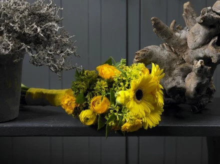 Styl klasyczny. Gerbera, jaskier i pieczennik. W tym klasycznym otoczeniu eleganckie kształty połączono z ciepłymi kolorami. Ten bukiet bardzo dobrze łączy oba te elementy. Różne kształty i żółty kolor kwiatów dodają pomieszczeniu ciepła. 