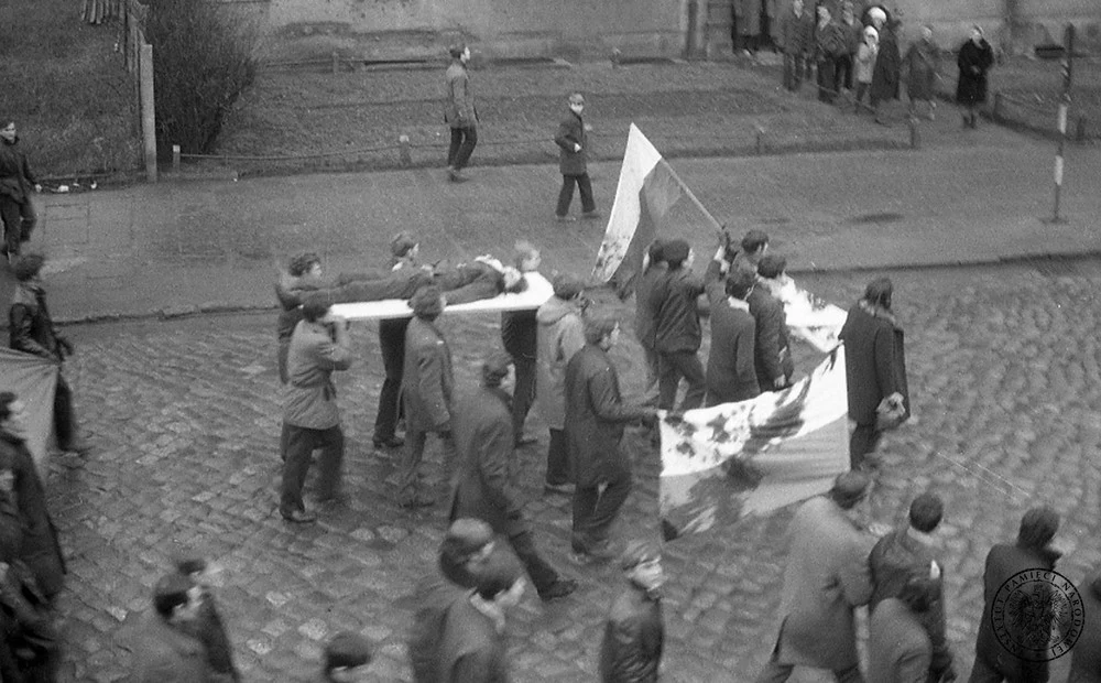 Protestujący maszerują ulicami Gdyni, niosąc na drzwiach ciało Zbigniewa Godlewskiego (znanego też jako Janek Wiśniewski) - robotnika zastrzelonego 17 grudnia 1970 r. W chwili śmierci miał 18 lat. Obok ciała - zakrwawione flagi narodowe Protestujący maszerują ulicami Gdyni, niosąc na drzwiach ciało Zbigniewa Godlewskiego (znanego też jako Janek Wiśniewski) - robotnika zastrzelonego 17 grudnia 1970 r. W chwili śmierci miał 18 lat. Obok ciała - zakrwawione flagi narodowe