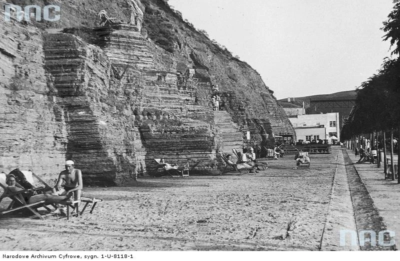 Zaleszczyki - fragment plaży nad Dniestrem. Widoczne schody wykute w skale oraz palający się kuracjusze. W oddali kiosk Polskiego Towarzystwa Księgarń Kolejowych RUCH Sp. z o.o. oraz fragment przęsła mostu kolejoweg