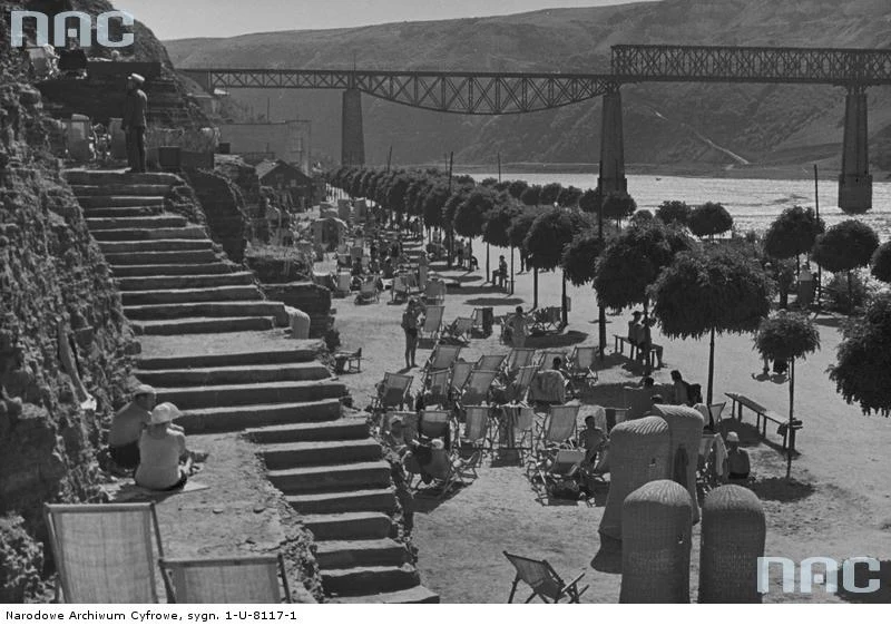 Zaleszczyki - fragment plaży z wykutym w skale zejściem. Widoczni kuracjusze opalający się na słońcu. Na drugim planie, na tle ściany jaru Dniestru widoczny odbudowany po zniszczeniach wojennych most kolejowy