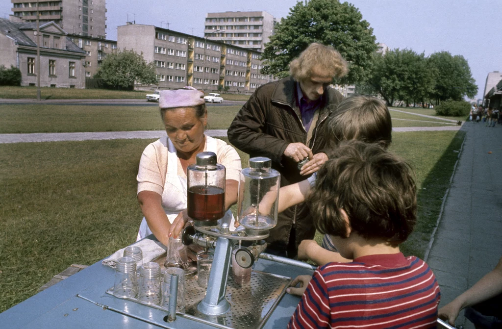 Warszawa, lata 70. Saturator z woda sodowa i sokiem stoi nieopodal pawilonów ze sklepami.