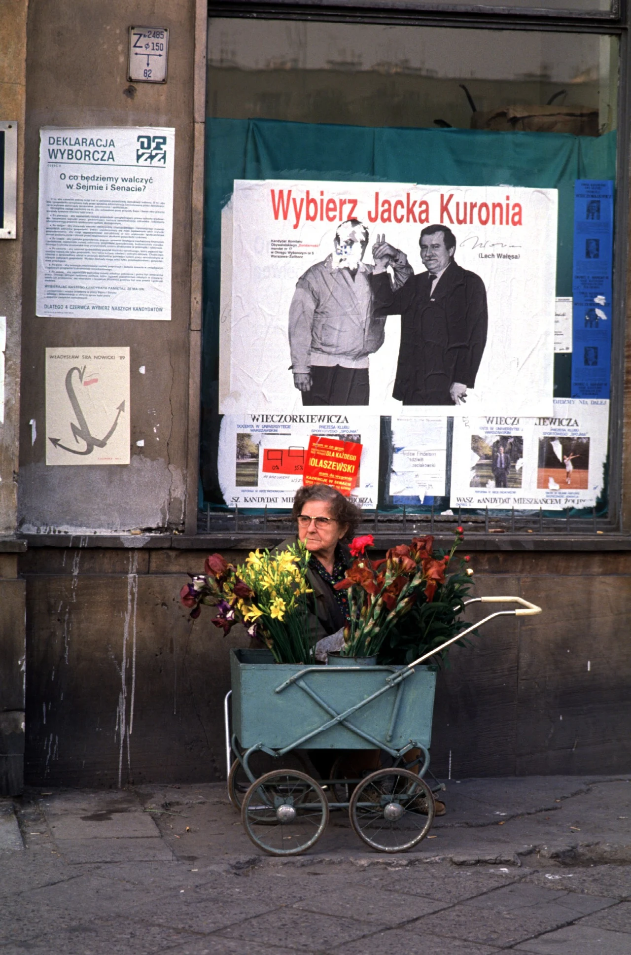 Starsza kobieta sprzedająca kwiaty na ulicy pod plakatem wyborczym Jacka Kuronia Starsza kobieta sprzedająca kwiaty na ulicy pod plakatem wyborczym Jacka Kuronia