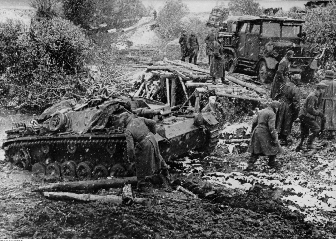Niemieckie działo samobieżne StuG III przejeżdża przez błotnisty teren na froncie wschodnim. Widoczna także ciężarówka jadąca po drewnianym moście