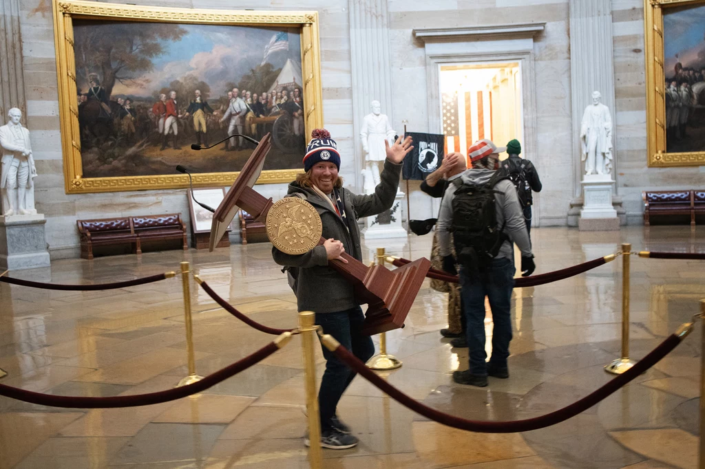36-letni Adam Johnson z Parrish na Florydzie został sfotografowany na ternie Kapitolu z ukradzioną mównicą Nancy Pelosi. Mężczyzna paradował w czapce z nazwiskiem Donalda Trumpa. Został aresztowany w piątek. Oskarżony jest o nielegalne wtargnięcie do budynku i kradzież własności federalnej. 36-letni Adam Johnson z Parrish na Florydzie został sfotografowany na ternie Kapitolu z ukradzioną mównicą Nancy Pelosi. Mężczyzna paradował w czapce z nazwiskiem Donalda Trumpa. Został aresztowany w piątek. Oskarżony jest o nielegalne wtargnięcie do budynku i kradzież własności federalnej.