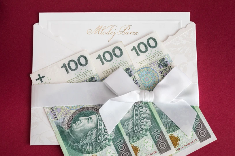 Pusta koperta? Taki gest może dotkliwie zaboleć Elegancka, jasna koperta ślubna z banknotami 100 zł, przewiązana białą wstążką na czerwonym tle.