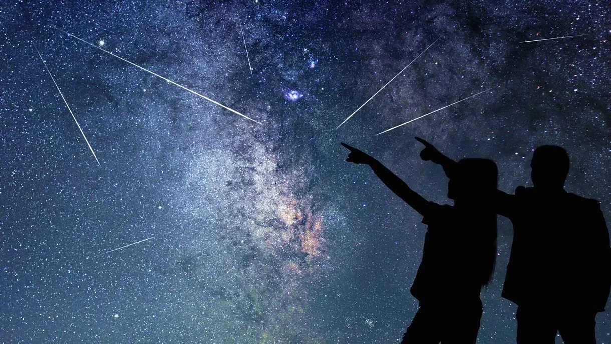 Niesamowity pokaz na niebie już dzisiaj. Nawet do 10 meteorów na godzinę Sylwetki dwóch osób na tle nocnego nieba z widocznymi gwiazdami, Drogą Mleczną i meteorami.