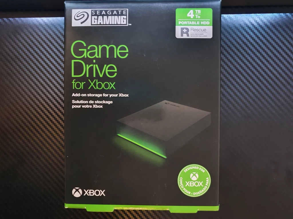 Dedykowany systemowi elektronicznej rozrywki Microsoftu dysk HDD z serii Game Drive firmy Seagate zapakowany został w prostokątne pudełko wielkości książki. Wewnątrz, między dobrze zabezpieczonymi plastikowymi profilami schowano właściwe urządzenie oraz kabel USB 3.2 Gen 1, skróconą instrukcję obsługi, dokument gwarancyjny i kartę z kluczem do usługi Xbox Game Pass Ultimate o ważności jednego miesiąca. Tradycyjny dysk talerzowy zamknięto w eleganckiej obudowie o minimalistycznym stylu z czarnego tworzywa sztucznego o wymiarach 12,25 cm x 8,1 cm x 2,15 cm. Urządzenie nie jest ciężkie, waga wynosi 266 g, z łatwością można się z nim przemieszczać, w czym pomaga kształt i zastosowanie matowego wykończenia, dzięki któremu na powierzchni nie odbijają się ślady palców. Dedykowany systemowi elektronicznej rozrywki Microsoftu dysk HDD z serii Game Drive firmy Seagate zapakowany został w prostokątne pudełko wielkości książki. Wewnątrz, między dobrze zabezpieczonymi plastikowymi profilami schowano właściwe urządzenie oraz kabel USB 3.2 Gen 1, skróconą instrukcję obsługi, dokument gwarancyjny i kartę z kluczem do usługi Xbox Game Pass Ultimate o ważności jednego miesiąca. Tradycyjny dysk talerzowy zamknięto w eleganckiej obudowie o minimalistycznym stylu z czarnego tworzywa sztucznego o wymiarach 12,25 cm x 8,1 cm x 2,15 cm. Urządzenie nie jest ciężkie, waga wynosi 266 g, z łatwością można się z nim przemieszczać, w czym pomaga kształt i zastosowanie matowego wykończenia, dzięki któremu na powierzchni nie odbijają się ślady palców.