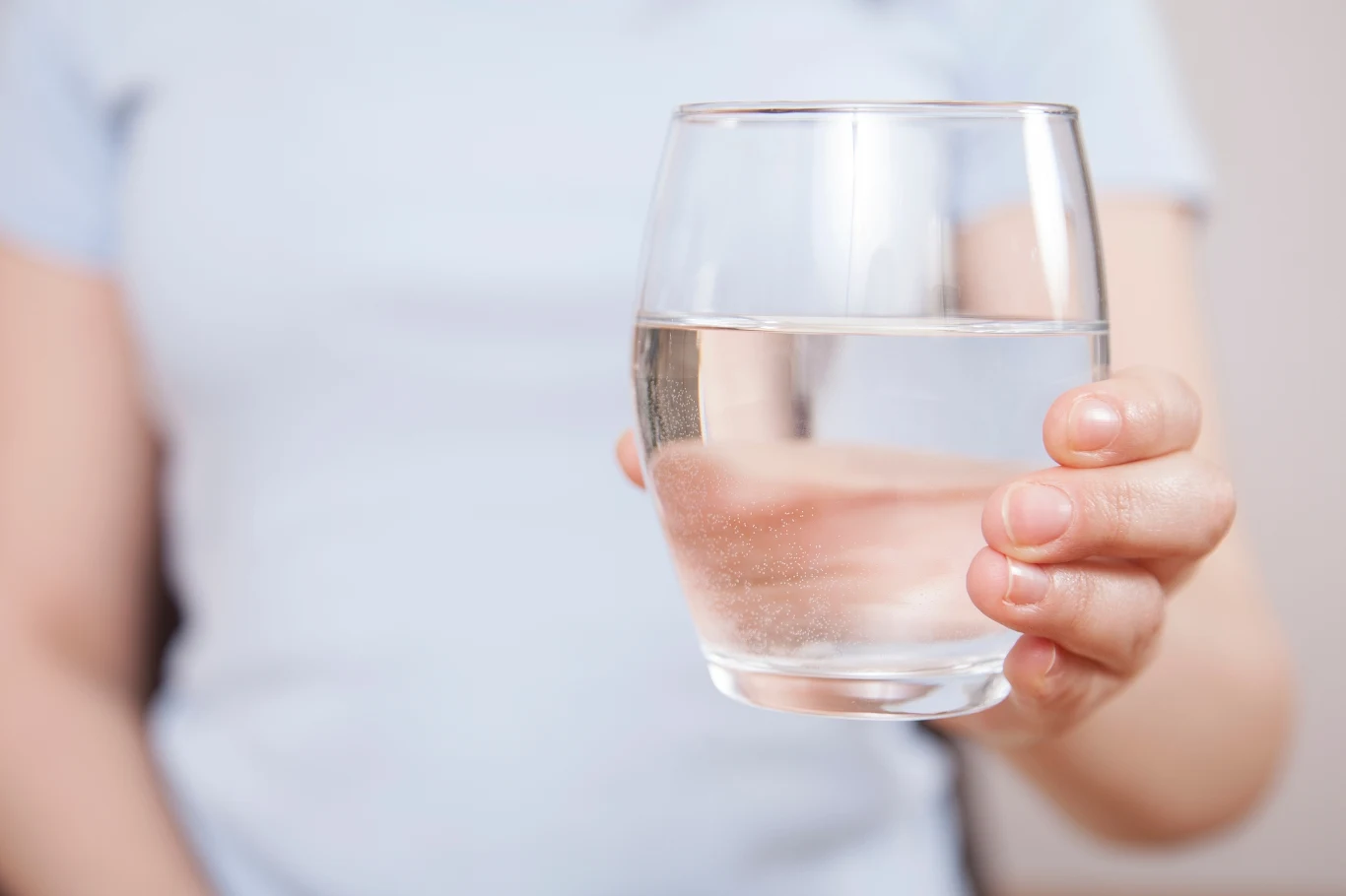 Jaką wodę naprawdę warto pić? Wybór nie zawsze jest tak oczywisty Dłoń trzymająca szklankę z wodą, widoczna część sylwetki osoby na tle jasnej koszulki.