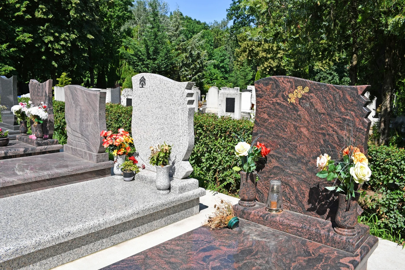 Ile kosztują nagrobki z granitu? Szereg nagrobków z polerowanego granitu z kwiatami i zniczami na cmentarzu otoczonym zielenią w słoneczny dzień
