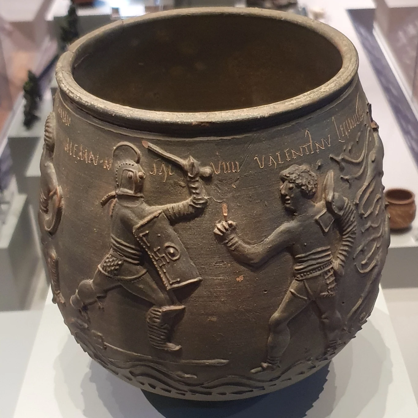 Memnon i Valentinus to zainscenizowane imiona. Ten pierwszy był w Rzymie utożsamiany z wieloma bardzo sławnymi, czarnoskórymi gladiatorami o wyjątkowych umiejętnościach. Nazwa pochodziła od legendarnego bohatera wojen trojańskich. Obraz na wazie przedstawia moment, gdy Memnon ma zadać ostateczny cios, gdy Valentinus podnosi palec do góry na znak poddania Memnon i Valentinus to zainscenizowane imiona. Ten pierwszy był w Rzymie utożsamiany z wieloma bardzo sławnymi, czarnoskórymi gladiatorami o wyjątkowych umiejętnościach. Nazwa pochodziła od legendarnego bohatera wojen trojańskich. Obraz na wazie przedstawia moment, gdy Memnon ma zadać ostateczny cios, gdy Valentinus podnosi palec do góry na znak poddania