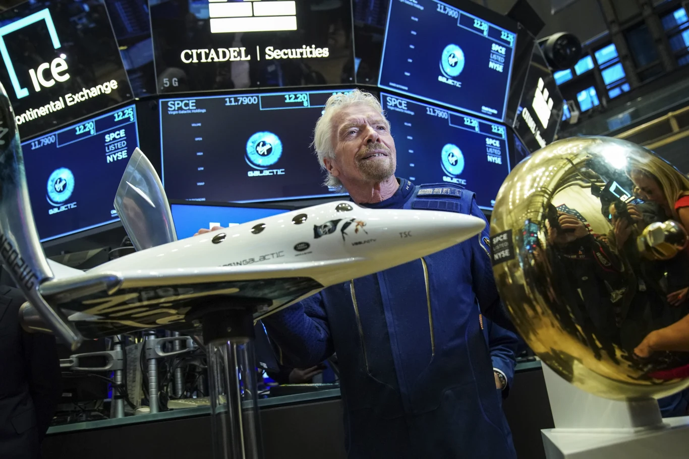 Richard Branson, założyciel Virgin Galactic z makietami statków kosmicznych i planet, na tle logo Virgin Group.
