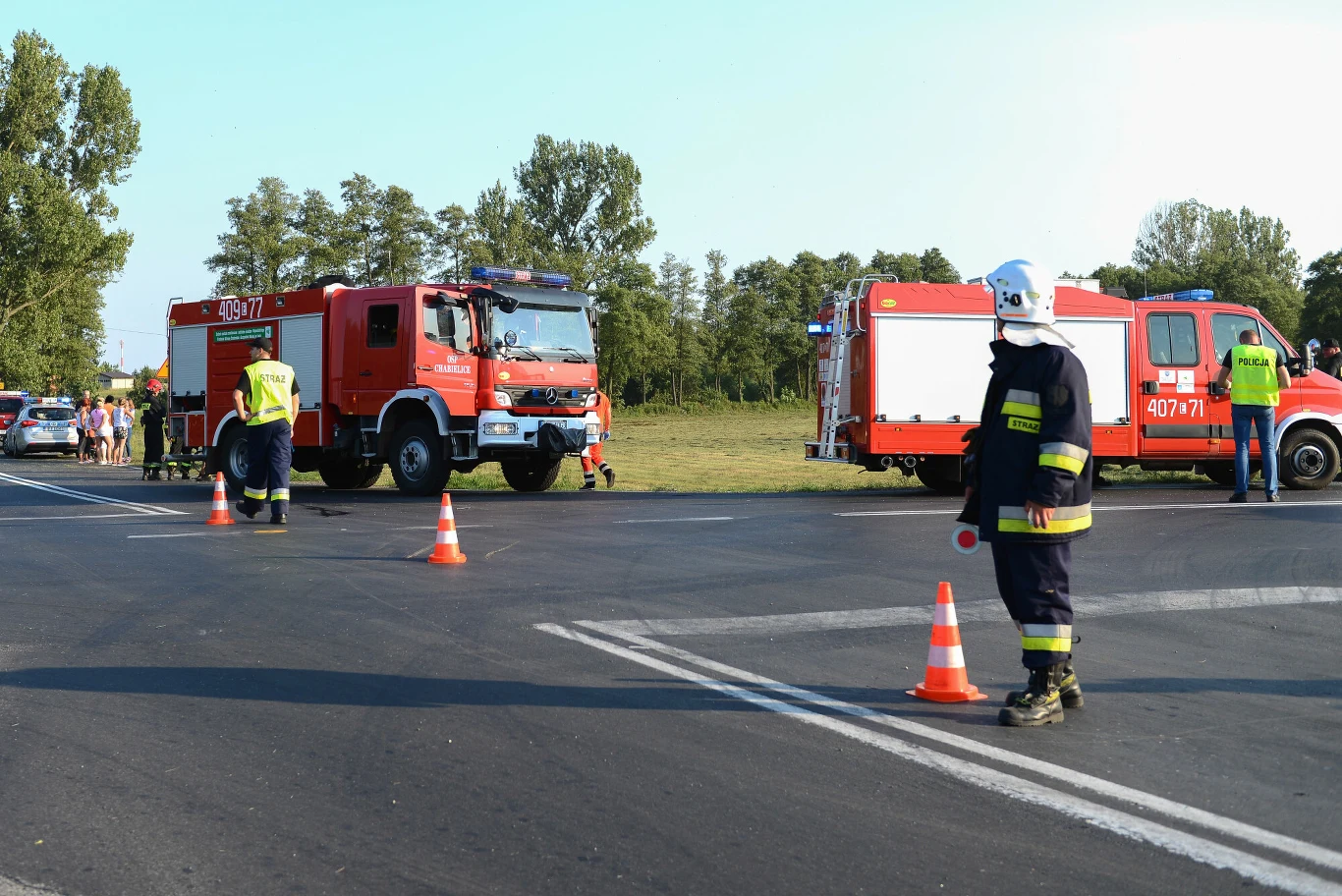 Poziomy znak P-12 "linia bezwzględnego zatrzymania" to gruby, biały pas prostopadły do ruchu pojazdów na danej drodze. Na zdjęciu widoczna tuż za stojącym najbliżej strażakiem.