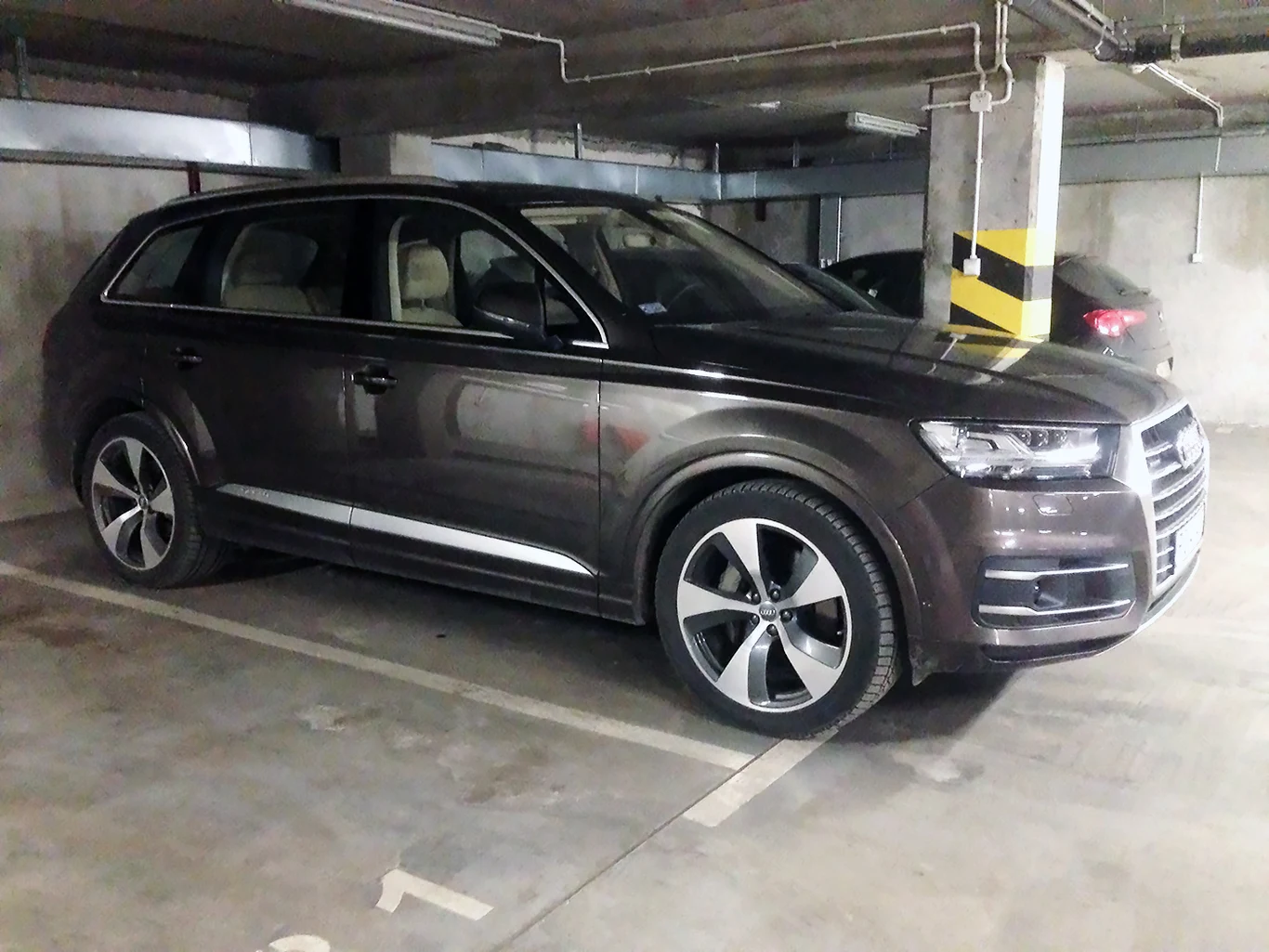 Samochody rosną, miejsca postojowe - nie. Carspereading staje się palącym problemem w centrach miast. Brązowe Audi Q7 SUV zaparkowane w podziemnym garażu, widoczne duże felgi, betonowe ściany i żółto-czarne słupy.