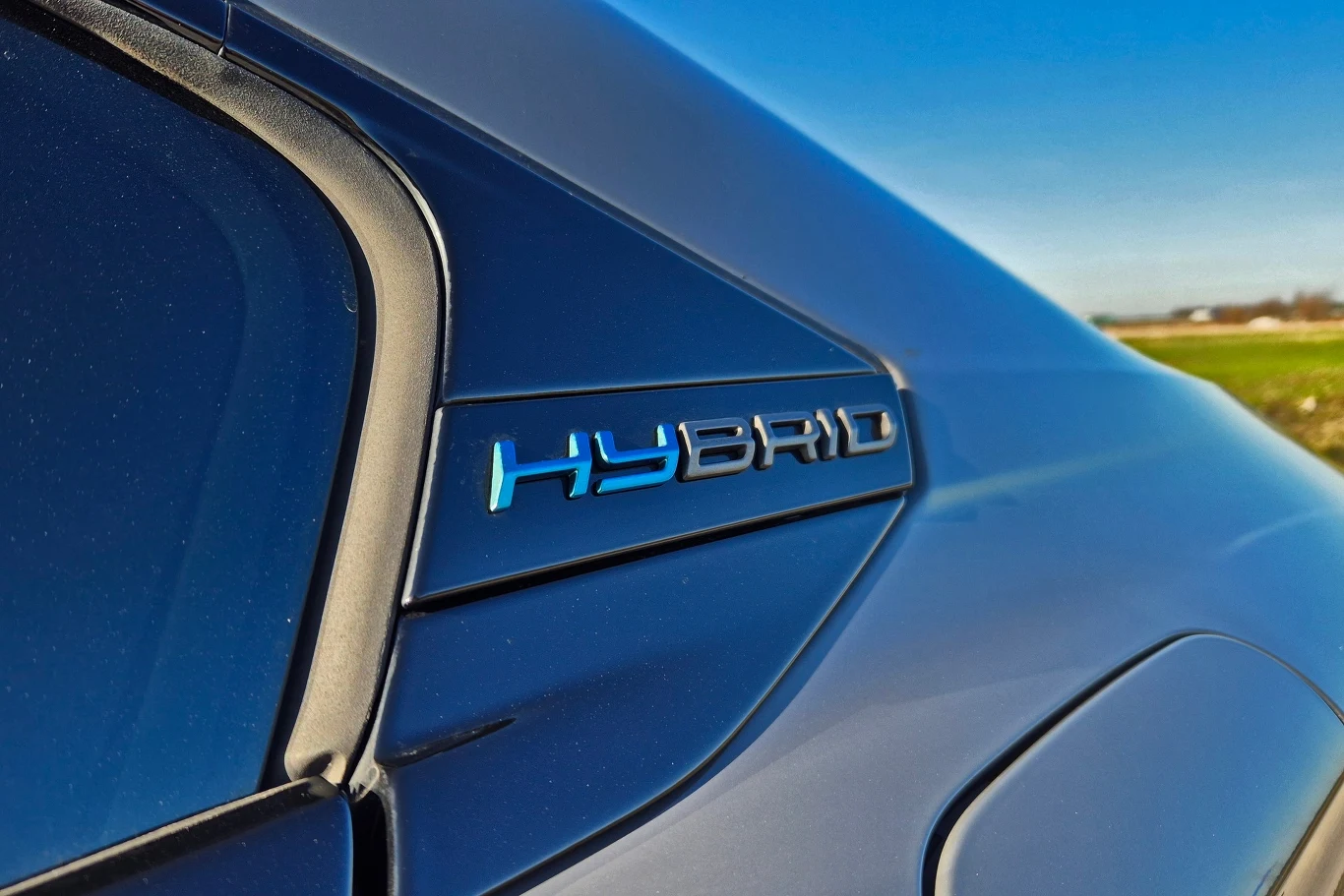 Hybrydy plug-in i kosztowne naprawy. Dlaczego PHEV tyle kosztują? Logo samochodu hybrydowego z napisem 'HYBRID' na niebieskiej, błyszczącej karoserii.