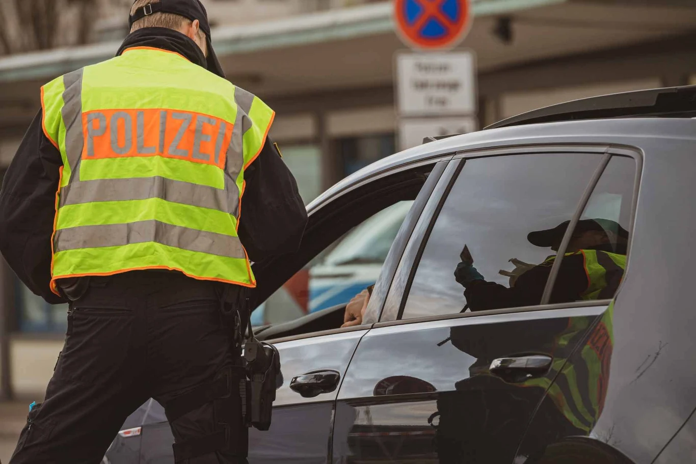 Czy za brak obowiązkowego wyposażenia auta za granicą można dostać mandat? Policjant w kamizelce odblaskowej rozmawia z kierowcą czarnego auta zatrzymanego przy krawężniku w mieście.