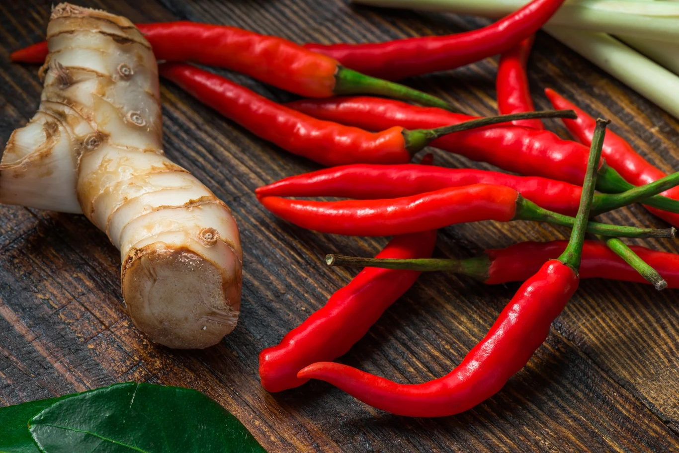 Chilli zawiera kapsaicynę, a w połączeniu z innymi przyprawami działa silnie przeciwzapalnie. Czerwone papryczki chili, galangal, liście i łodygi trawy cytrynowej na ciemnym drewnie, wyraziste barwy przypraw.