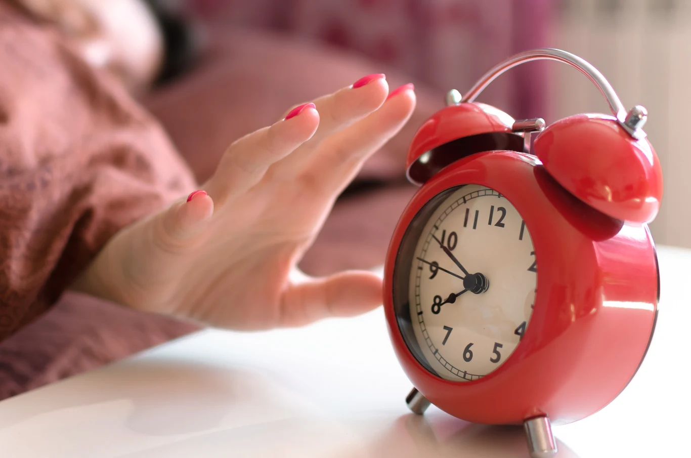 Kiedy w tym roku nastąpi zmiana czasu z letniego na zimowy? Czerwony budzik na stoliku nocnym pokazuje prawie siódmą, dłoń sięga po wyłącznik, w tle łóżko z osobą.