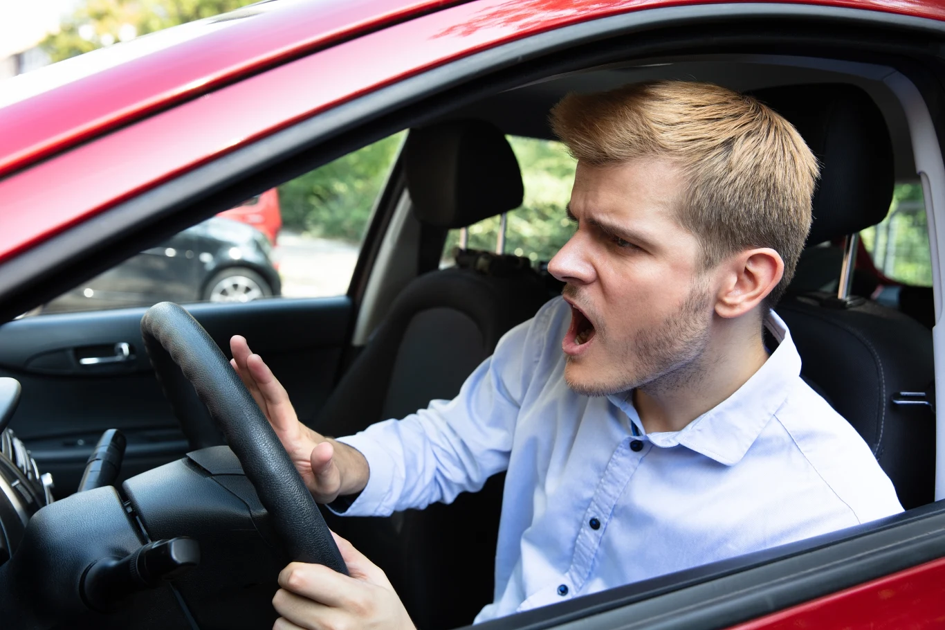 Policyjne skrzynki "Stop agresji drogowej" pękają w szwach. Rekordowy wzrost liczby zgłoszeń Mężczyzna w czerwonym samochodzie gestykuluje ręką przy kierownicy; ilustracja wzrostu zgłoszeń o agresję drogową.