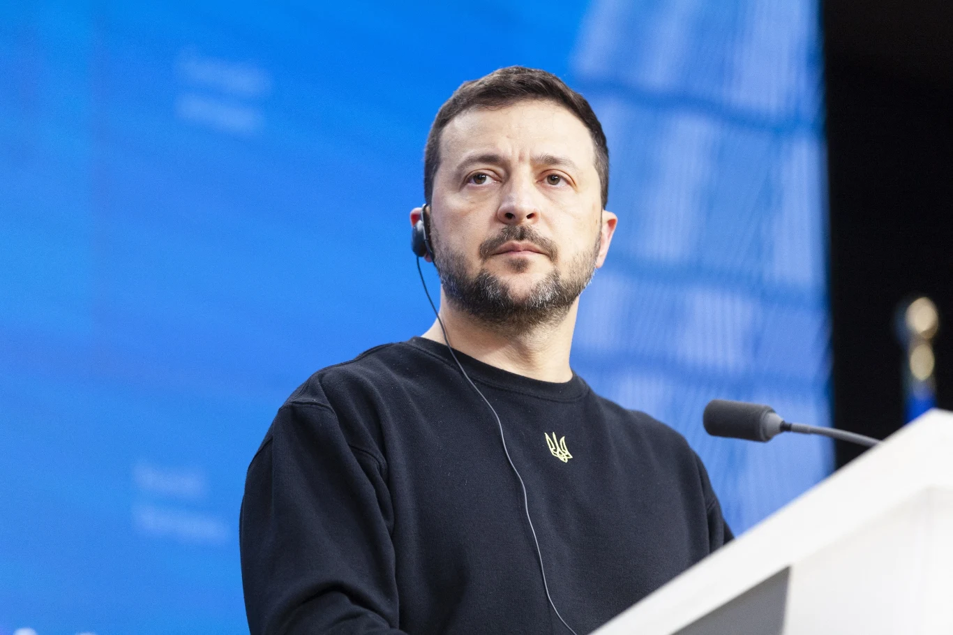 Prezydent Ukrainy Wołodymyr Zełenski Wołodymyr Zełenski przemawia do mikrofonu w ciemnej bluzie na niebieskim tle