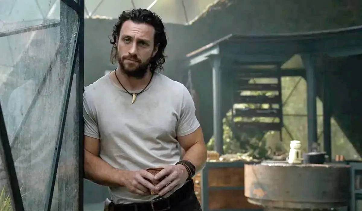 Aaron Taylor-Johnson w scenie z filmu "Kraven Łowca" Aaron Taylor-Johnson jako Kraven Łowca oparty o metalową konstrukcję w szklarni, w tle rośliny i stół.